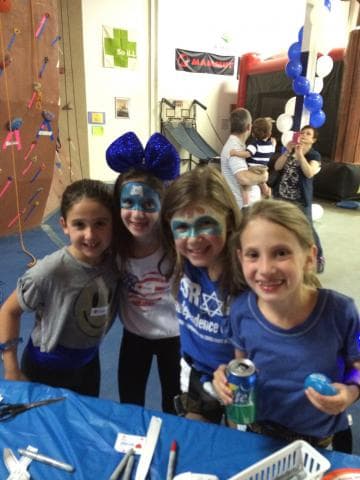 An image of four girls dressed up; some with their faces painted and another big a big blue bow on her head. They are smiling at the camera and embracing each other.