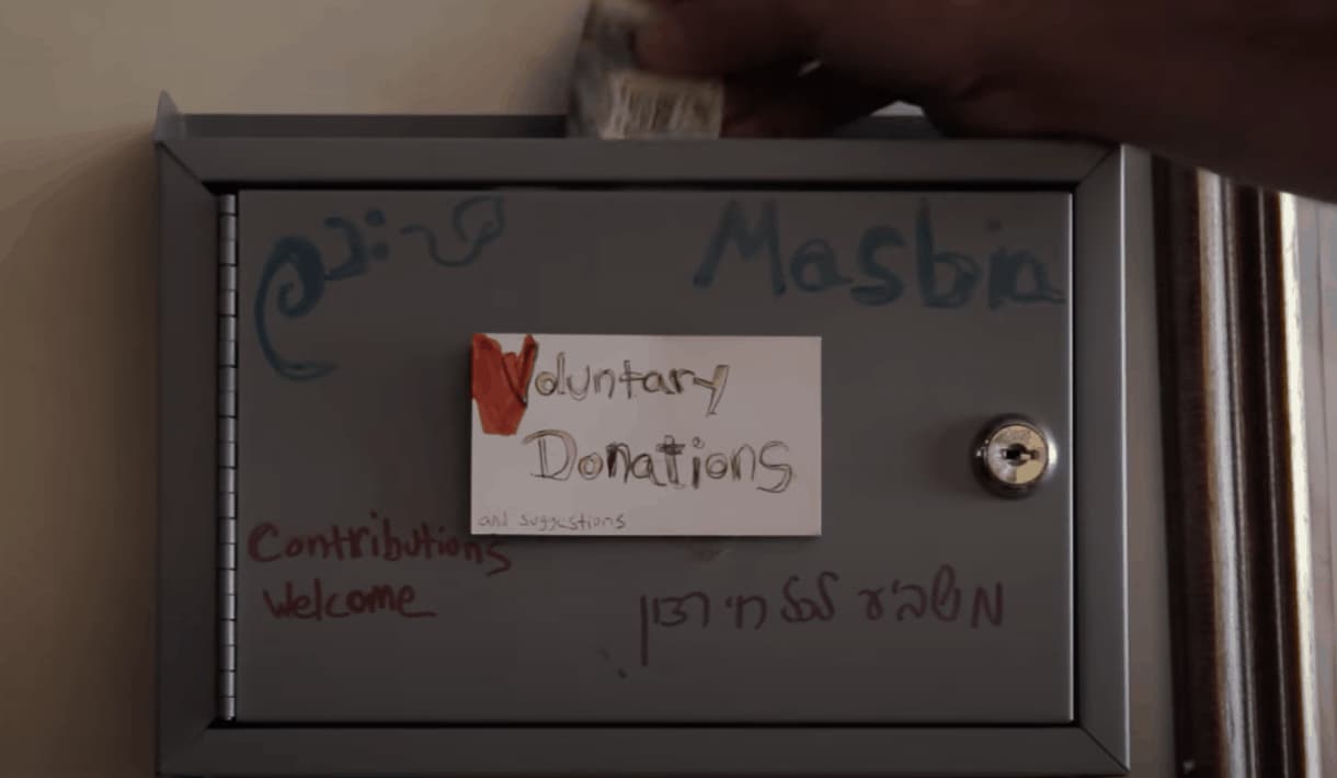A screenshot from a video showing a lockbox labeled "Masbia - Voluntary Donations - Contributions Welcome" with a hand putting a dollar into the box.
