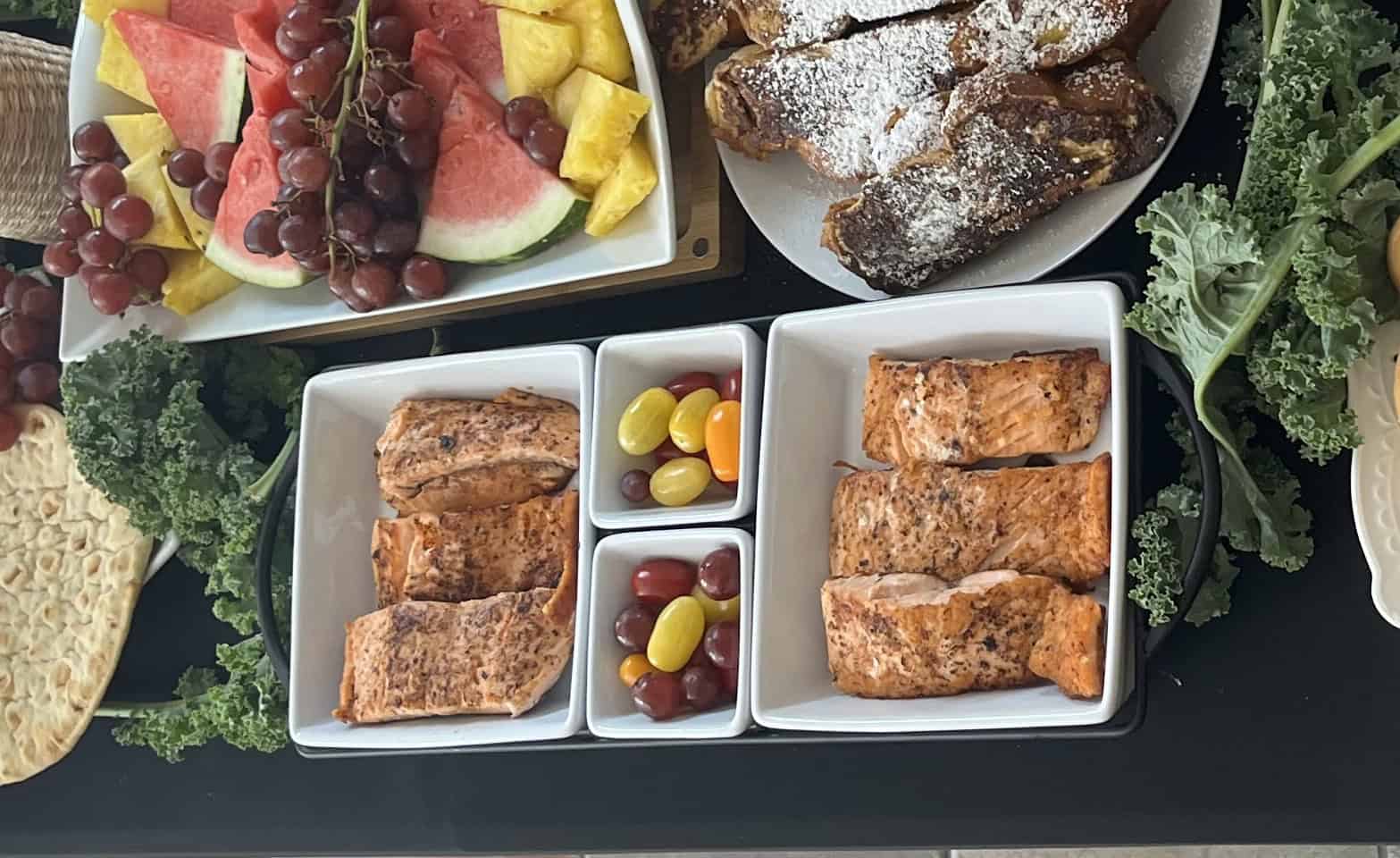 A photo of several plates of grilled salmon on a table with other plates with fruit, bread, and olives.
